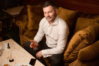 Restoranda lüks bir kanepede otururken bir bardak pahalı konyakla takılan zarif, ciddi bir adam.