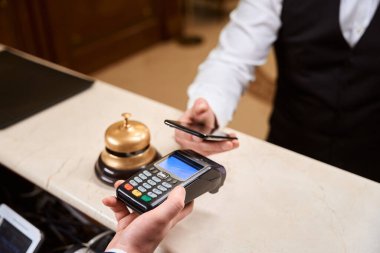Otel resepsiyonunda yönetici kredi kartı makinesini tutarken cep telefonu başvurusuyla temas kurmadan ödeme yapan erkek misafiri kapat.