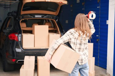Bayan, kutularla dolu bir arabanın bagajının yanında duruyor. Elinde büyük bir karton kutu ve bant tabancası var.