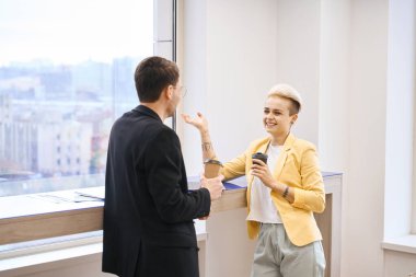 Arkadaş canlısı iş arkadaşları kahve içerken ve kahve molası sırasında konuşurken, pencerenin yanındaki tezgaha yaslanırken, dinlenirken