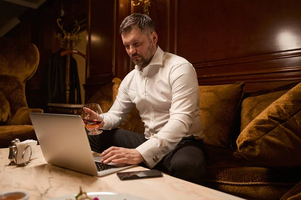 在餐馆里 一个人坐在沙发上喝威士忌时 一个英俊的留着胡子的男人在笔记本电脑上工作 — 图库照片