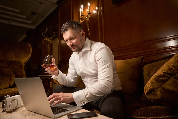 在餐馆里 大男人坐在沙发上 一个人坐着喝着一大杯白兰地 一边用笔记本电脑 — 图库照片
