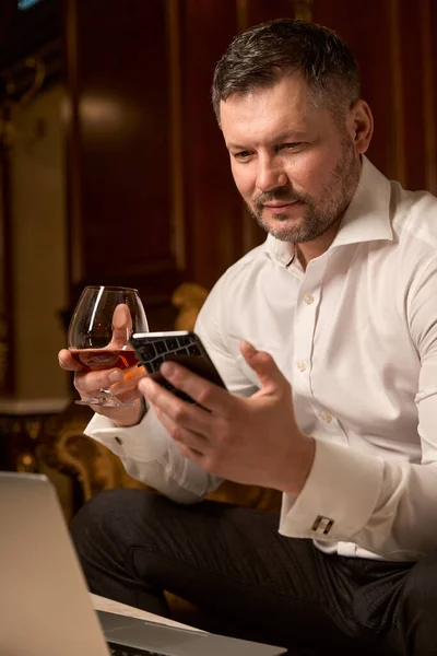 豪华餐厅的沙发上 一个留着胡子的英俊男人一边用智能手机一边喝着一杯白兰地 一边放松自己 — 图库照片