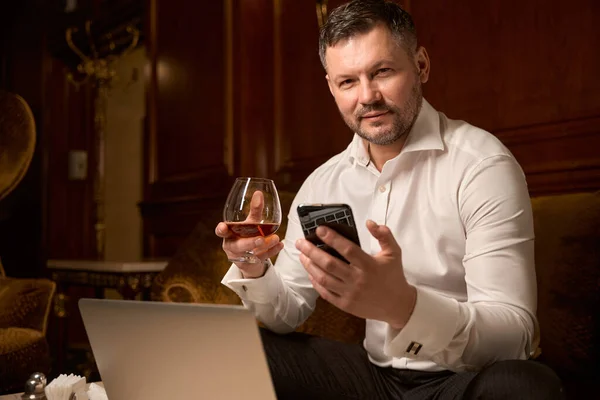 一个自信而严肃的男人一边在餐馆的沙发上喝一杯白兰地 一边用智能手机给妻子发短信 — 图库照片