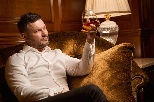 Elégant Homme Sérieux Examinant Verre Avec Whisky Cher Tout Reposant — Photo