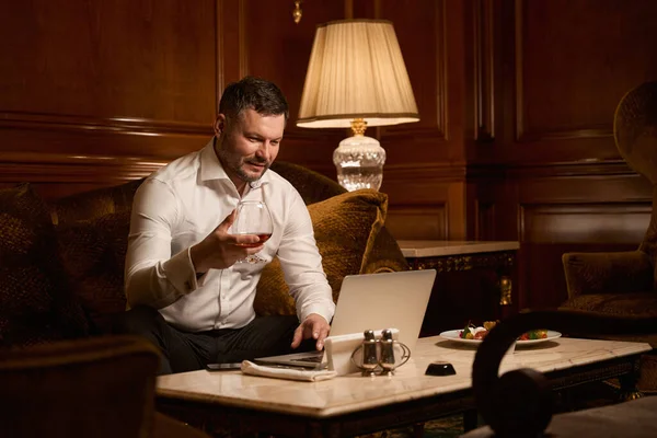 一个英俊的男人坐在餐馆的沙发上喝酒 一边用手提电脑发短信 — 图库照片