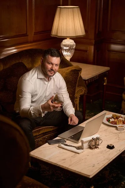 レストランでソフトソファの上でラップトップを使用している間魅力的な男性のゲストは 新しい高価なブランデーを試飲 — ストック写真