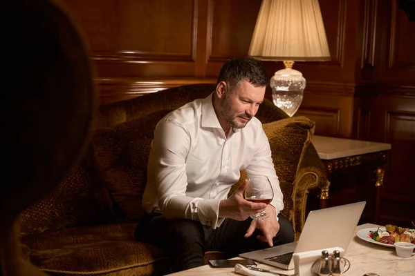 在豪华酒店的沙发上 大商人一边拿着杯子 一边在笔记本电脑上与伴侣聊天 — 图库照片