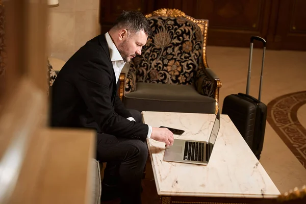 在酒店大堂 长相漂亮的男人坐在扶手椅上休息 并在靠近行李的桌子边的手提电脑上浏览一些东西 — 图库照片