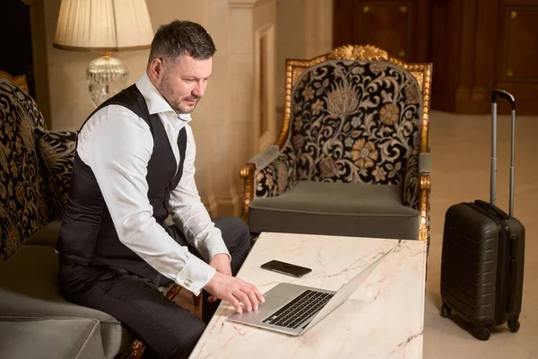 在酒店大堂靠近行李的桌子边 一位英俊而严肃的商人在笔记本电脑上工作 — 图库照片