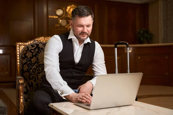 在酒店大堂 穿着正式西装的英俊优雅的商人坐在扶手椅上 在行李箱旁边的桌子旁使用笔记本电脑 — 图库照片
