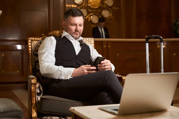 양복을 사업가가 스마트폰을 로비에 앉아서 호텔을 시간을 기다리고 있습니다 — 스톡 사진