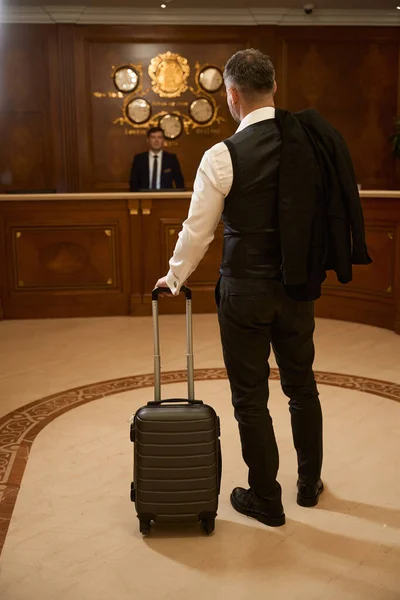 身着黑色西服的商人背着行李站在酒店大堂 而男性接待员则坐在接待处的后面 — 图库照片