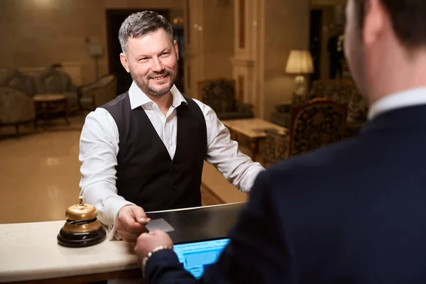 ホテルのロビーで受付にサービスを支払うためのクレジットカードを与える正式な訴訟でハンサムな笑顔の男 — ストック写真