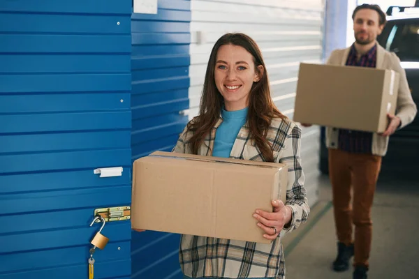 微笑的女士拿着一个大盒子靠近储藏室敞开的门 在仓库里 在后台 一个人拿着一个盒子 里面装的是汽车里的东西 — 图库照片