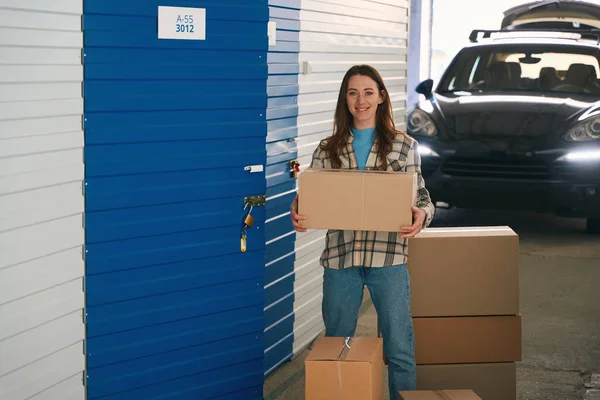 微笑的女士拿着一个大盒子靠近储藏室敞开的门 在仓库里 后面站着一辆开着行李箱的车 — 图库照片