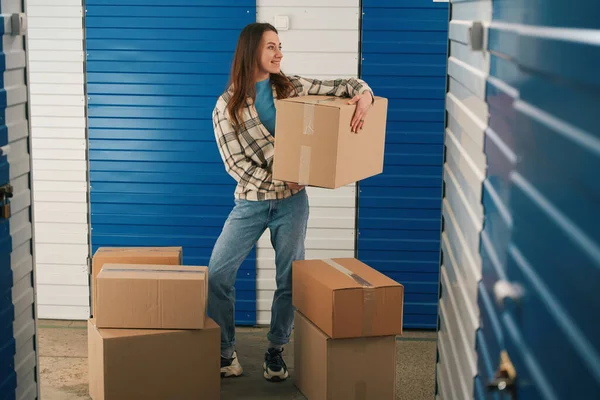 年轻的女士手里拿着一个大纸板箱 站在仓库里 周围有许多箱子 — 图库照片