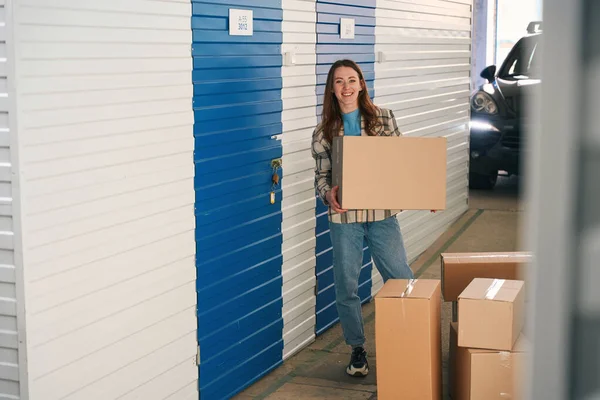 Young woman is standing near the storage room in the warehouse and holding a big cardboard box