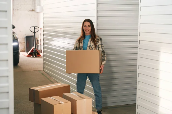 幸せな女性は大きな段ボール箱を保持し 周りの多くのボックスと倉庫に立っている — ストック写真