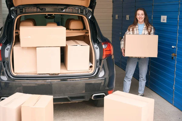 若い女性が倉庫に箱と大きな段ボール箱を持って車のオープントランクの近くに立っている — ストック写真