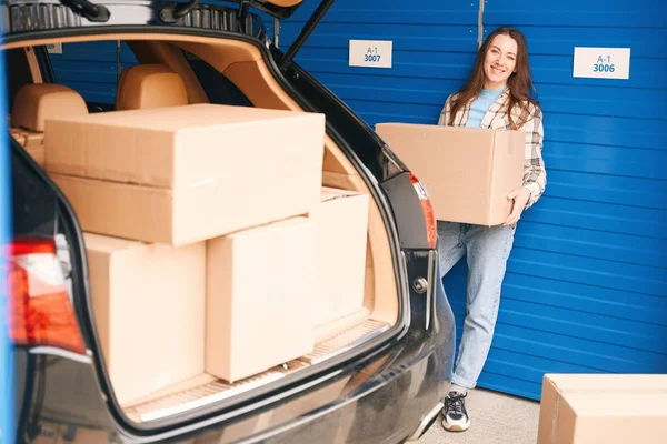 若い女性が倉庫に箱と大きな段ボール箱を持って車のオープントランクの近くに立っている — ストック写真