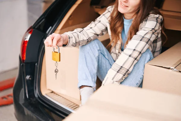 笑顔の女性が倉庫に鍵を手に車のトランクに座っている 移動のためのものと段ボール箱はトランクにあり 車の隣にあります — ストック写真