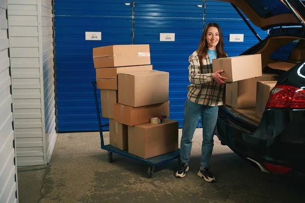 微笑的女人把纸板箱放进汽车后备箱 放进装有自动贮存器的仓库 — 图库照片