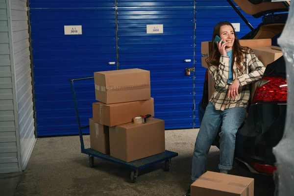 微笑的女士拿着手机打电话 坐在仓库里的汽车后备箱里 装有物品的纸板箱在月台卡车上 在汽车后备箱里 — 图库照片