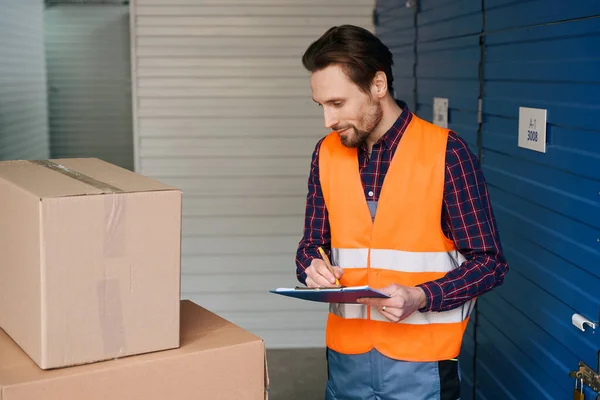 Young guy is standing near cardboard boxes and writing notes with tablets for writing in storage warehouse