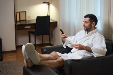 Orta yaşlı bir otel misafiri bir fincan kahveyle dinlenme alanında oturuyor, cep telefonuyla iletişim kuruyor.