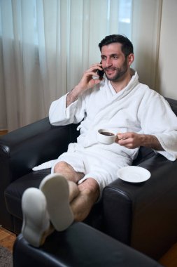 Tıraşsız, bornozlu bir erkek rahat deri koltukta oturuyor, elinde bir fincan kahveyle cep telefonuyla konuşuyor.