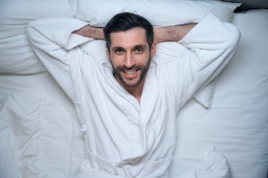 Mutlu dinlenmiş bir adam otel odasında uzanıyor, üzerinde rahat bir bornoz var.