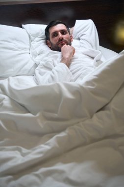 Hüzünlü bir adam otel odasında yumuşak yastıklarda yatıyor, bornozlu.