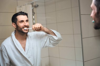 Gülümseyen orta yaşlı bir adam banyoda dişlerini fırçalıyor, beyaz bornozlu.