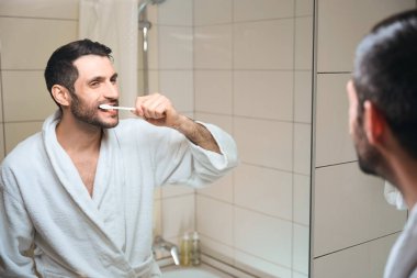 Gülümseyen orta yaşlı esmer, dişlerini aynanın önünde fırçalıyor, beyaz bornoz giyiyor.