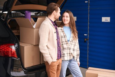 Gülümseyen kadın ve adam bir depoda karton kutularla açık bir arabanın bagajında birbirlerine bakıyorlar.