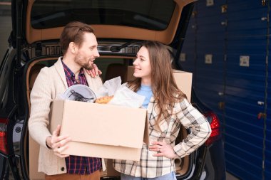 Gülümseyen kadın ve adam bir depoda açık bir arabanın bagajında birbirlerine bakıyorlar. Adamın elinde büyük bir karton kutu var.