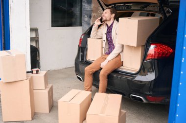 Mutlu adam bir depoda oturuyor ve bir arabanın bagajını arıyor. İçinde taşınacak eşyalar olan karton kutular bagajda ve arabanın yanında.