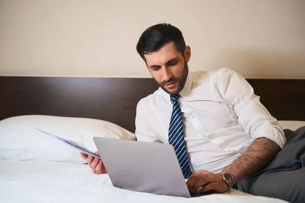 穿着工作服的未剃光头发的男人靠在床上 带着工作文件和笔记本电脑 — 图库照片