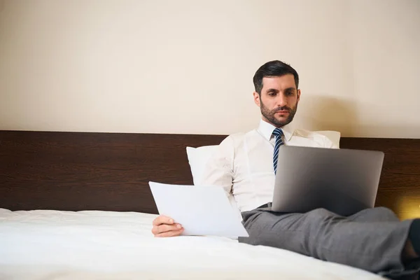 英俊的男人半坐在枕头上 躺在床上 手里拿着工作文件和笔记本电脑 — 图库照片