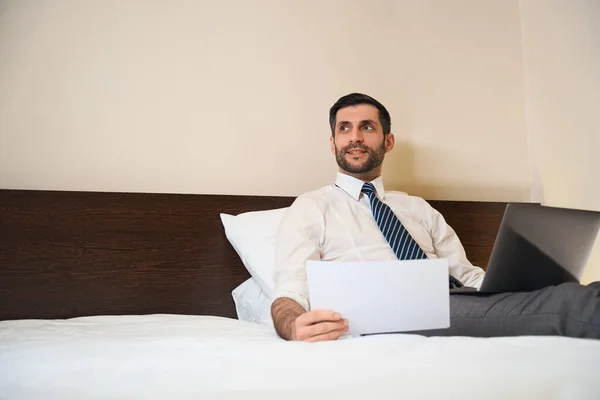 男人半坐在枕头上 靠在床上 带着工作文件和笔记本电脑 — 图库照片