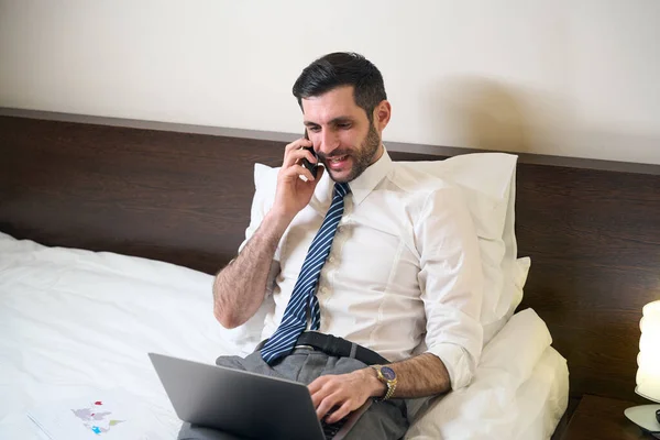 穿着办公室衣服的男人躺在一张大床上 他使用笔记本电脑和电话 — 图库照片