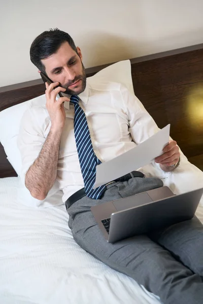 大規模なベッドの上にオフィス服の男性があり 彼は仕事文書を勉強し 電話で通信します — ストック写真