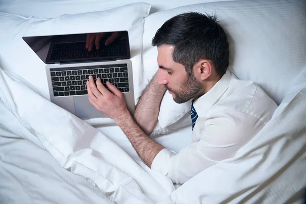 疲れたビジネスマンは ラップトップで作業中に服を脱がずに眠る 彼は快適なベッドに位置しています — ストック写真