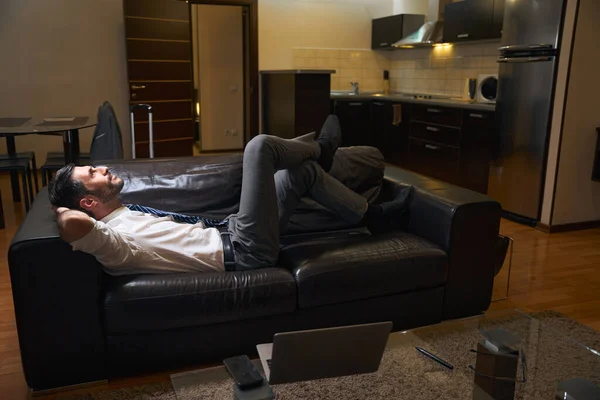 穿着办公室服装的经理躺在宾馆房间的皮沙发上 笔记本电脑和电话放在桌子上 — 图库照片