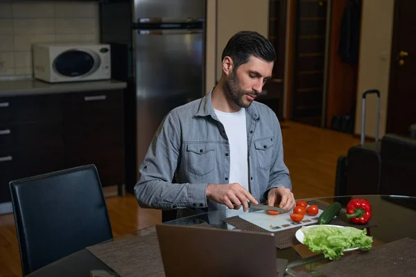 男人切西红柿当蔬菜沙拉 旁边的桌子是笔记本电脑 — 图库照片