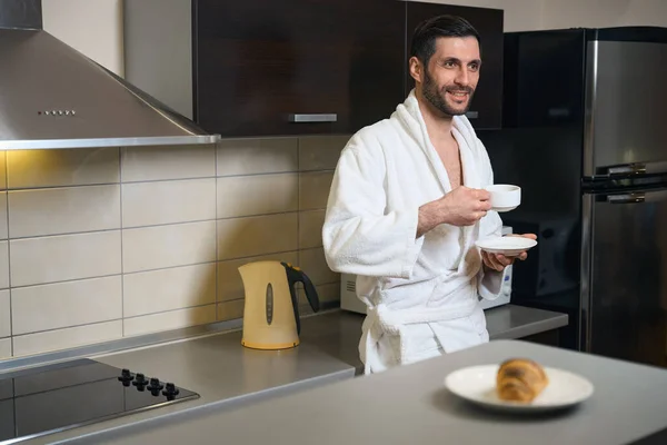 穿着浴衣的未刮胡子男子站在厨房区 手里拿着一杯咖啡 厨房的小玩意就在附近 — 图库照片
