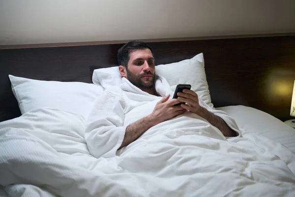 Hombre Cansado Está Descansando Cama Sobre Almohadas Suaves Tiene Teléfono — Foto de Stock