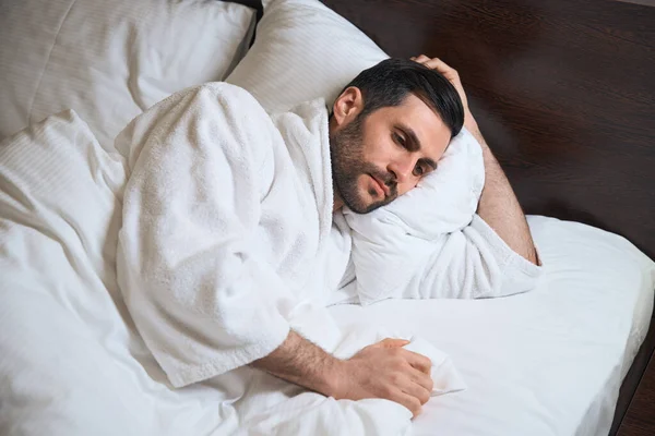 疲れたホテルのゲストは柔らかい枕の上のベッドの上に横になり 彼はバスローブにあります — ストック写真