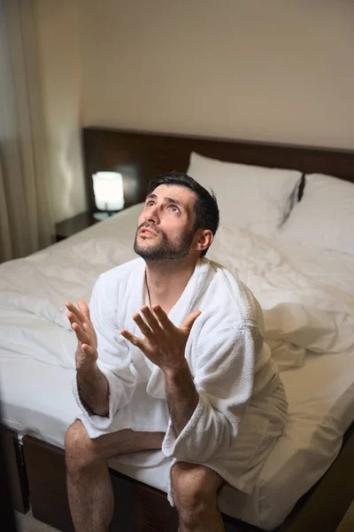 穿着浴衣的未剃光的男性坐在宾馆房间的一张大床上 他向天堂呼唤 — 图库照片